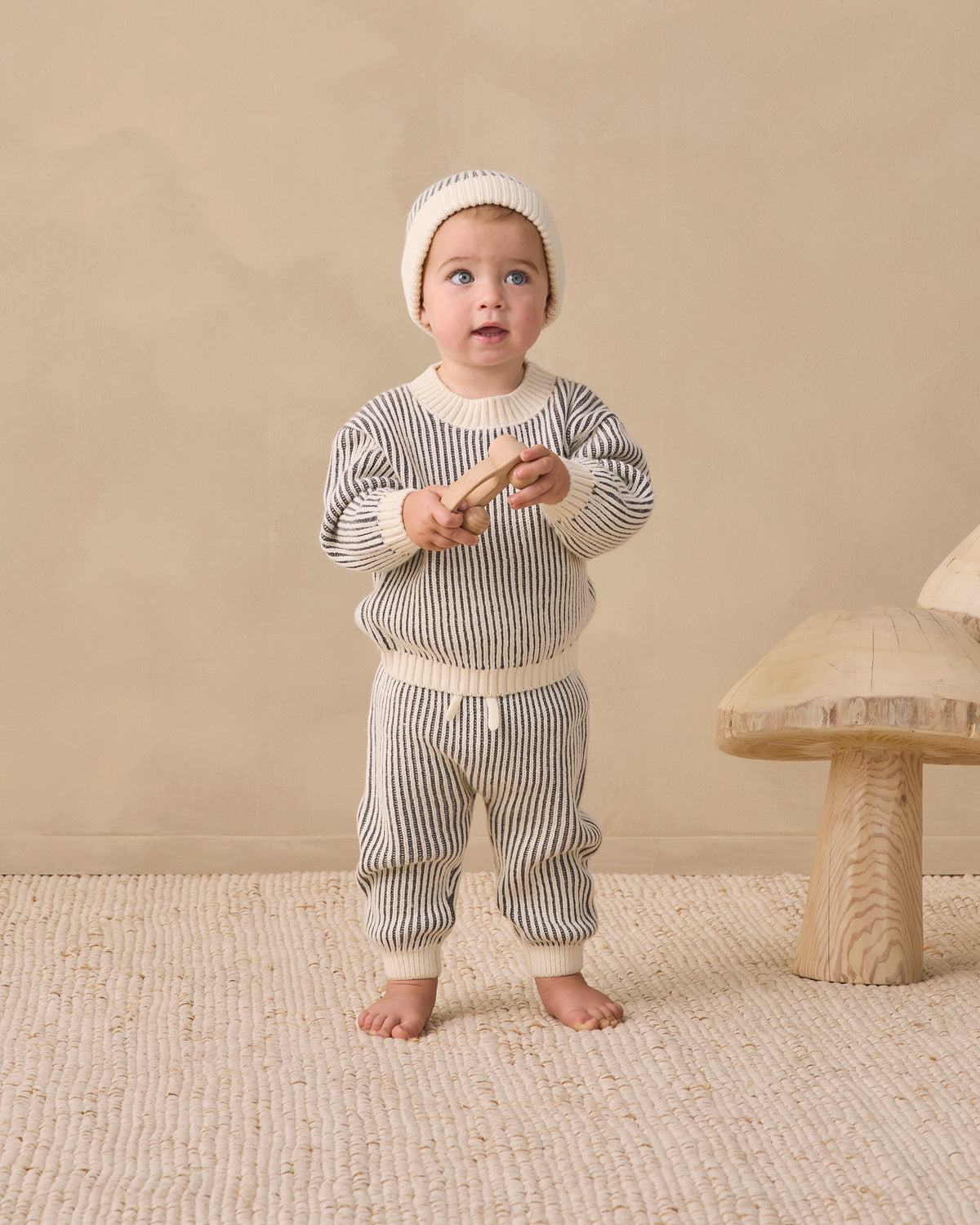 A baby standing barefoot on a textured beige rug, wearing a cream knit beanie and a matching cream and gray striped knit two-piece outfit. The background is a solid beige wall with a rustic wooden mushroom-shaped stool on the right.