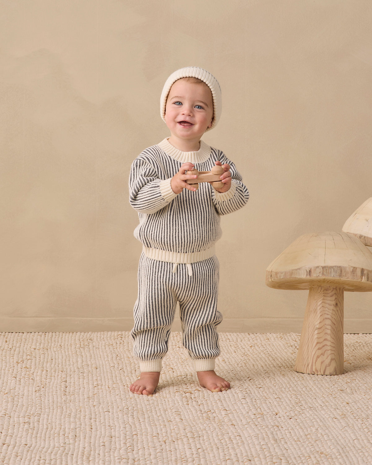 A baby wearing a cream knit two-piece outfit with thin gray vertical stripes, including a long-sleeve top and matching pants, standing barefoot on a textured beige rug next to a wooden mushroom toy.