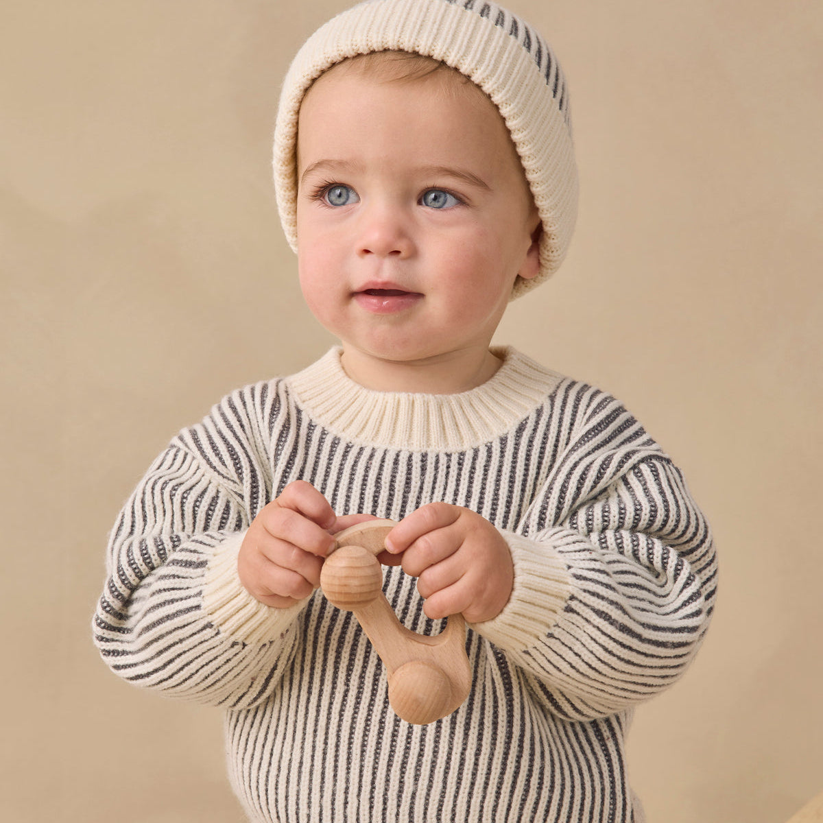 A baby wearing a cream and navy striped knit sweater and matching beanie hat, holding a wooden teething toy against a warm beige background.