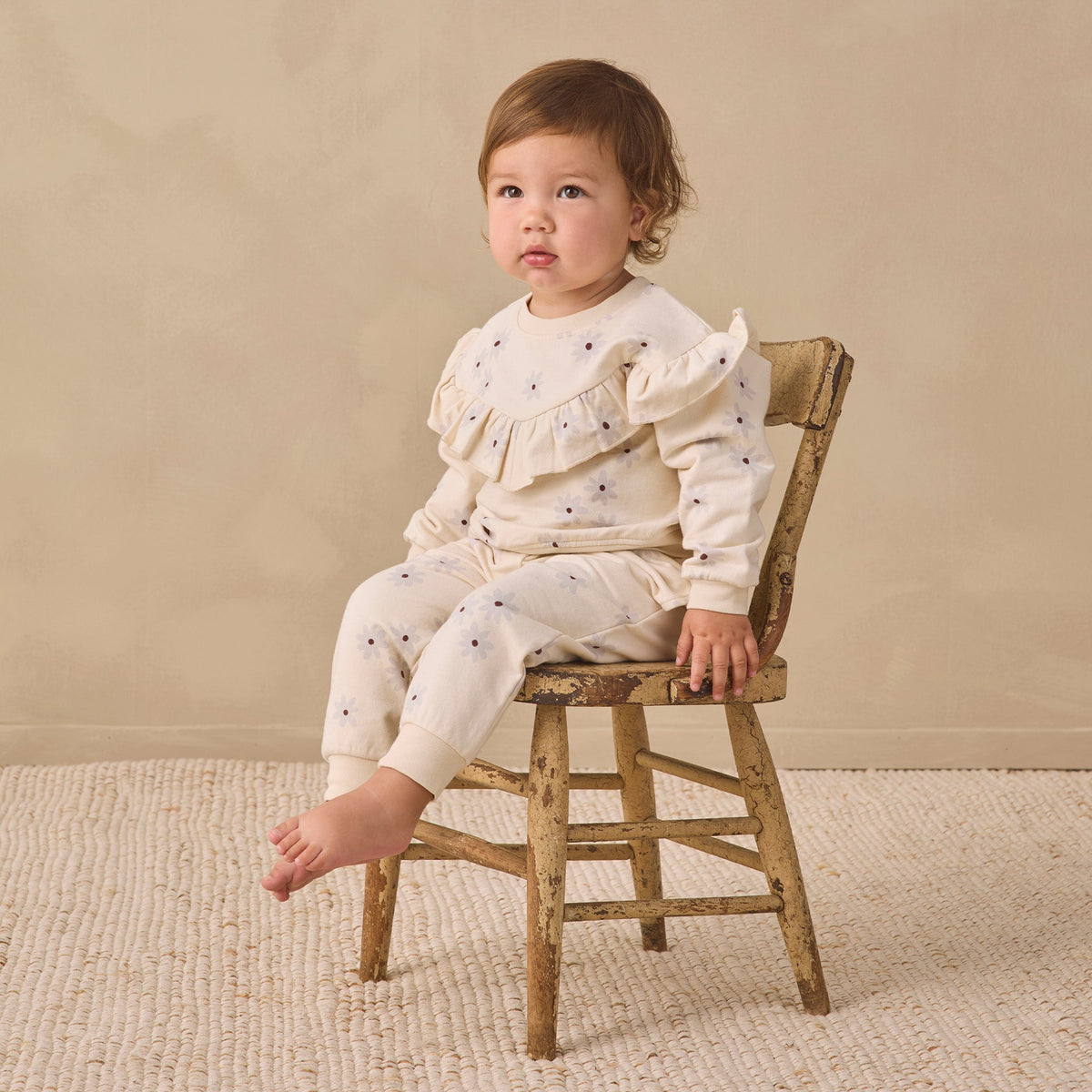 A toddler sits on a small rustic wooden chair wearing a cream ruffled long-sleeve top and matching pants with tiny dark blue star prints, set against a warm beige studio backdrop.
