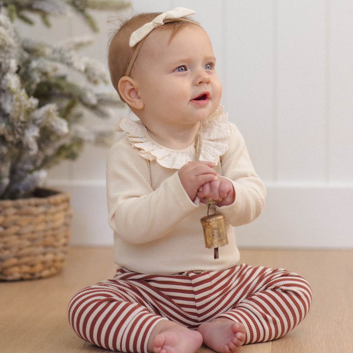 A baby sitting on a wooden floor, wearing a long-sleeve natural beige ruffled collar bodysuit and burgundy-and-white striped pants, holding a small brass bell while wearing a headband with a bow.