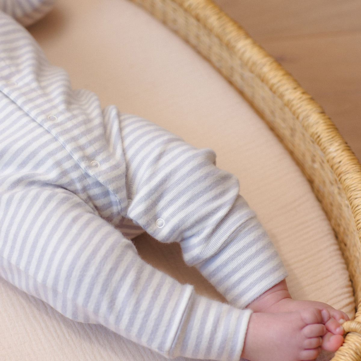 A close-up of a baby's legs and feet resting inside a round wicker basket, wearing a soft, organic cotton long-sleeve romper in a white and gray horizontal stripe pattern by Quincy Mae.