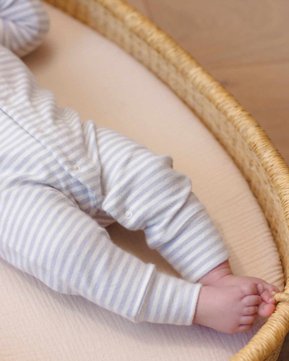 A close-up of a baby's legs and feet resting inside a round wicker basket, wearing a soft, organic cotton long-sleeve romper in a white and gray horizontal stripe pattern by Quincy Mae.