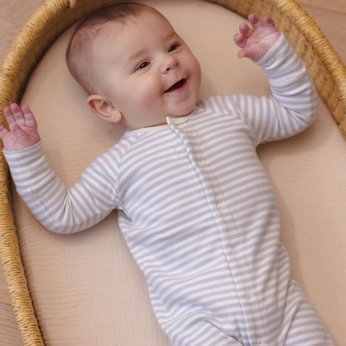 A smiling baby lying inside a round wicker basket, wearing a soft, organic cotton long-sleeve romper in a white and gray horizontal stripe pattern by Quincy Mae.