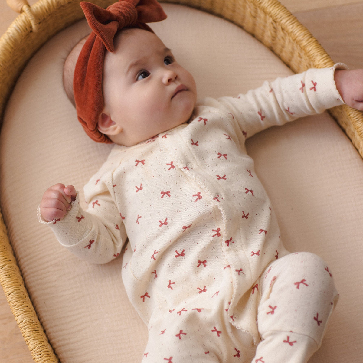 Baby wearing a redwood velour bow headband and a patterned cream-colored outfit, lying in a wicker basket.