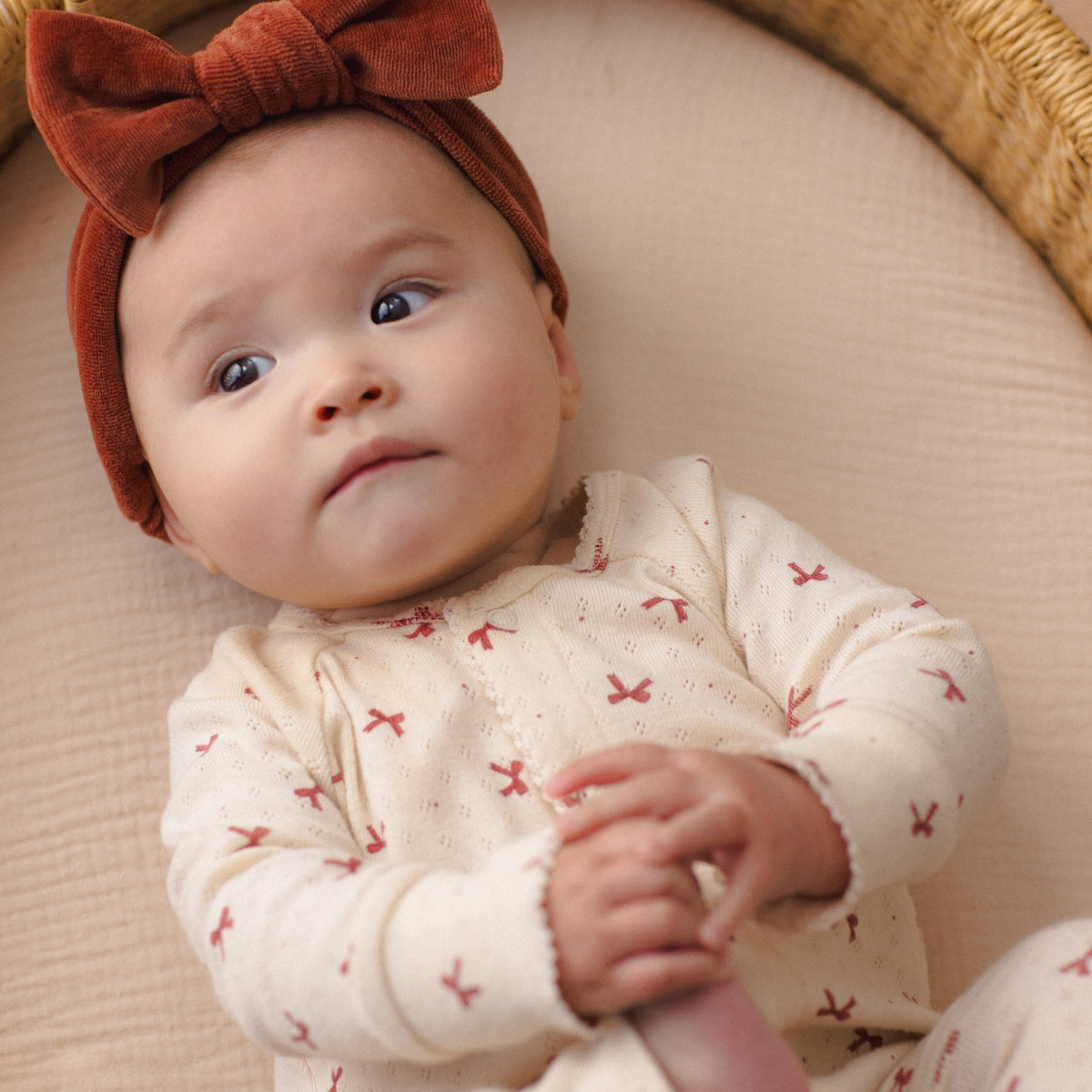 Baby wearing a redwood velour bow headband and a patterned cream-colored outfit, lying in a wicker basket.