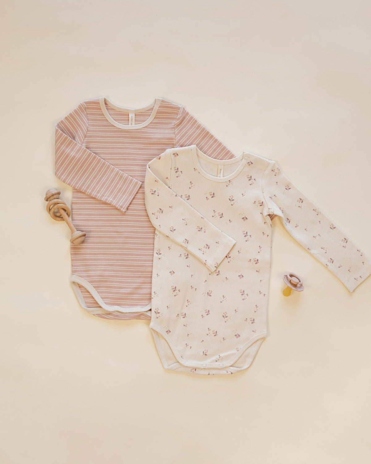 Two long-sleeve baby bodysuits laid flat on a warm cream background. The left bodysuit is light pink with thin white stripes, and the right bodysuit is off-white with a small scattered floral print.
