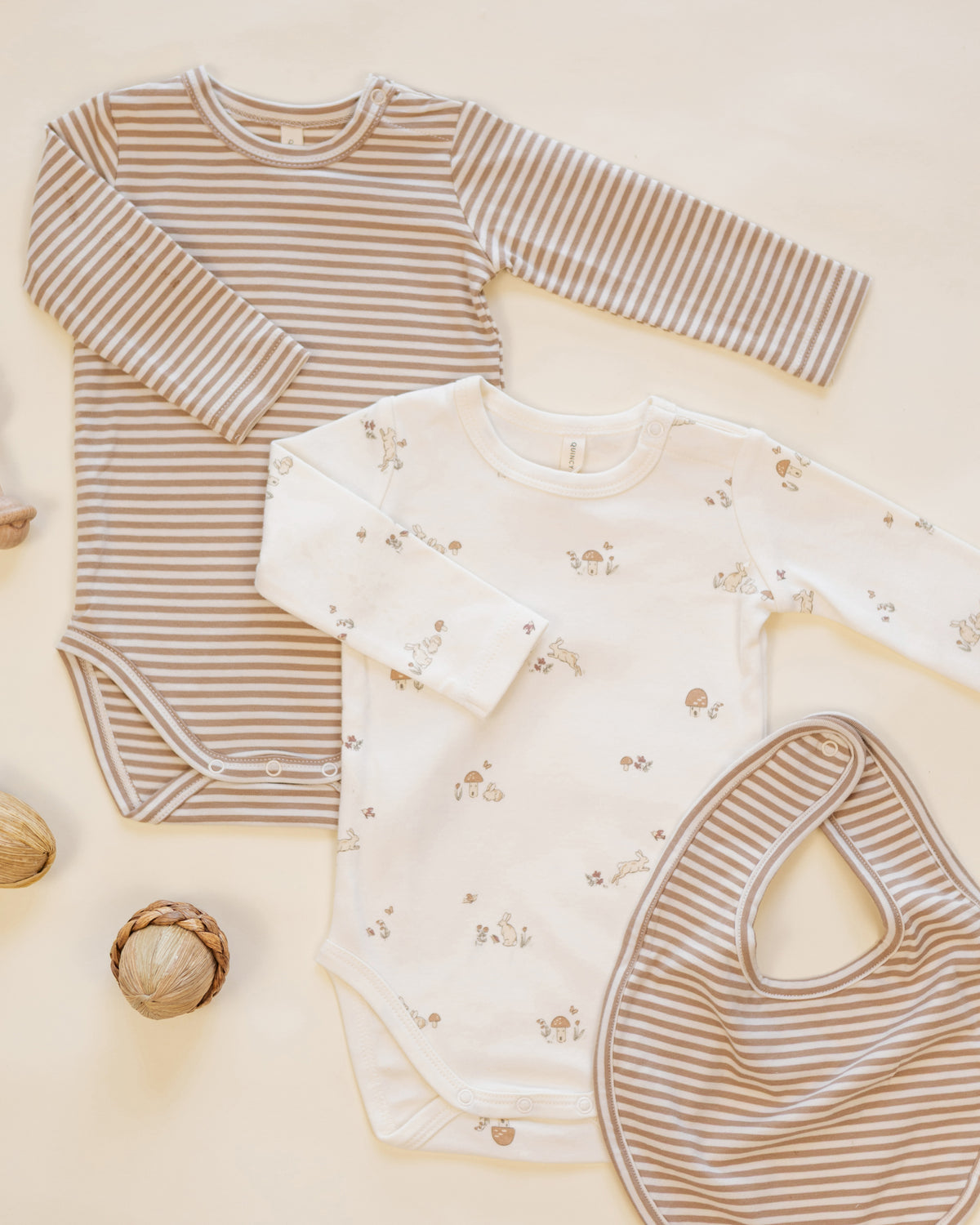 Three baby clothing items arranged in a flat lay on a warm cream background: a long-sleeve bodysuit with beige and white stripes, a long-sleeve bodysuit with a small floral print on cream, and a matching striped bib.