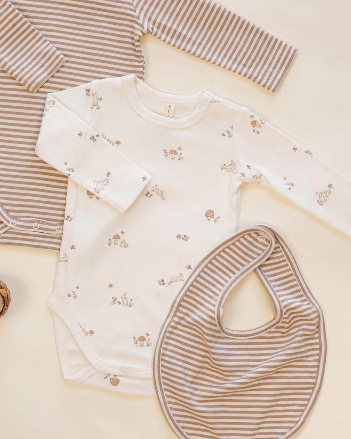 A baby clothing flat lay featuring a long-sleeve bodysuit in off-white with tiny brown animal prints, a matching striped long-sleeve top in beige and brown, and a striped bib.