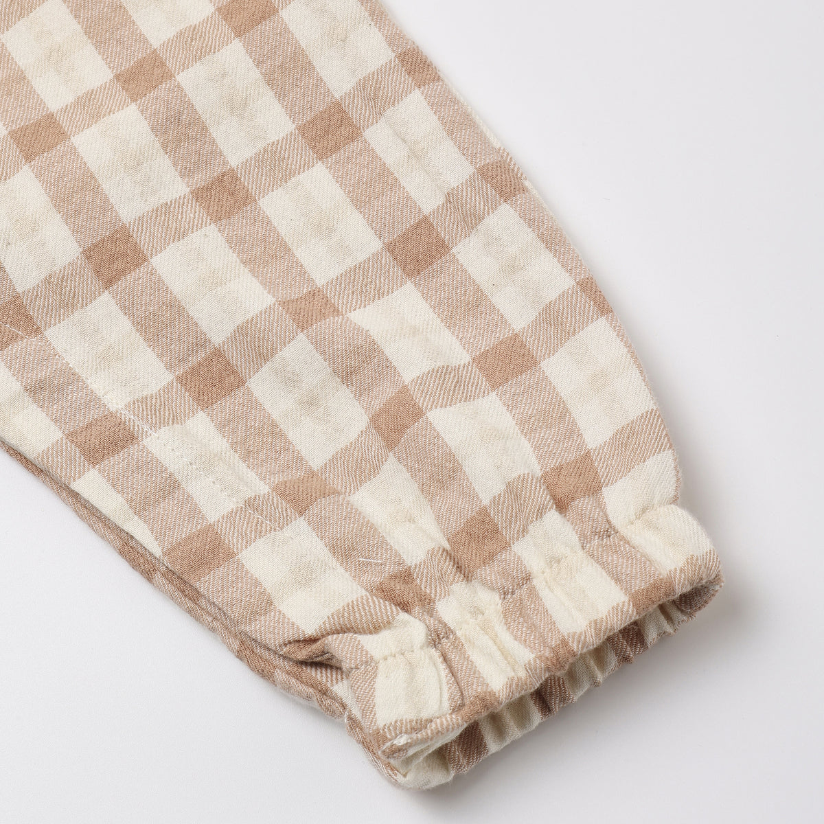 A close-up of a beige and off-white gingham checkered fabric item, showing a gathered elastic cuff on a light gray background.