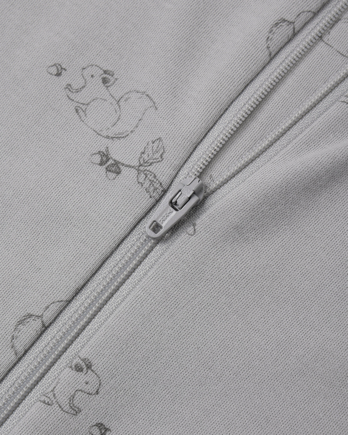 Close-up of a light gray zippered garment with a subtle repeating duck and leaf print, showing the zipper pull and fabric texture.