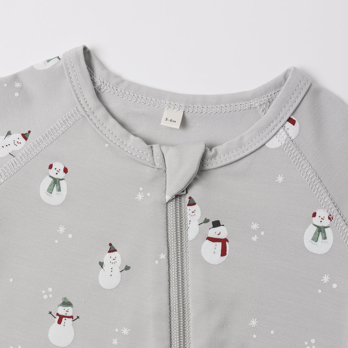 A close-up view of the upper chest and collar area of a light grey Quincy Mae bamboo zip footie, featuring an all-over wintery snowman print and a central zipper.