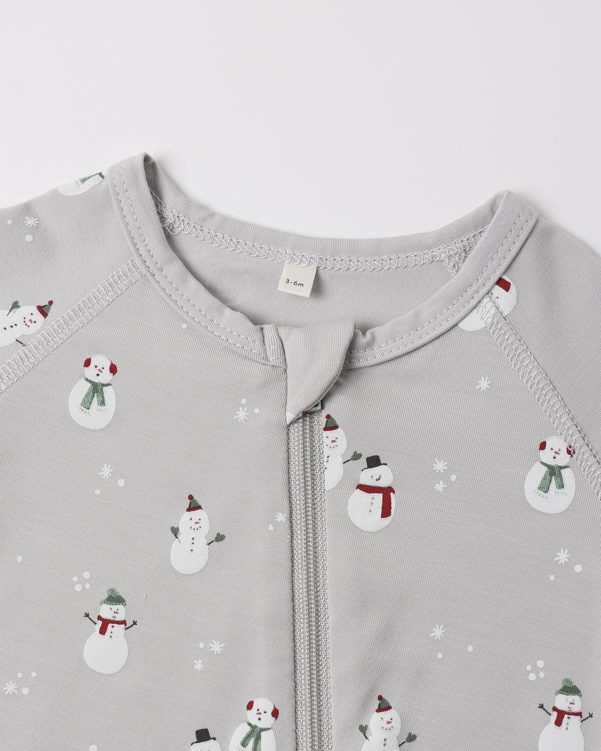 A close-up view of the upper chest and collar area of a light grey Quincy Mae bamboo zip footie, featuring an all-over wintery snowman print and a central zipper.