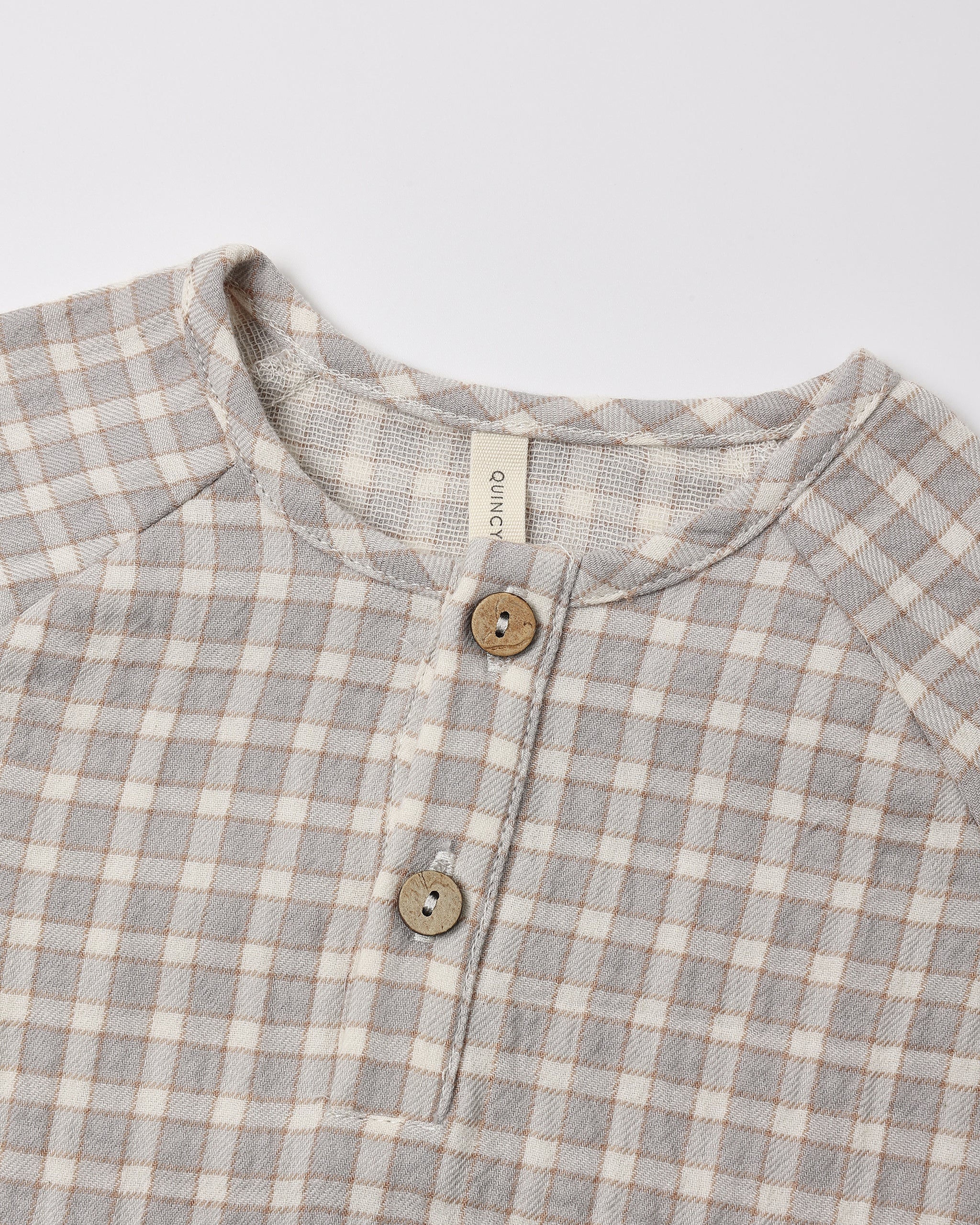 Close-up of a children's long-sleeve henley shirt with a light gray and beige gingham check pattern, featuring a button placket with wooden buttons and a visible neck label reading 'gugu'.