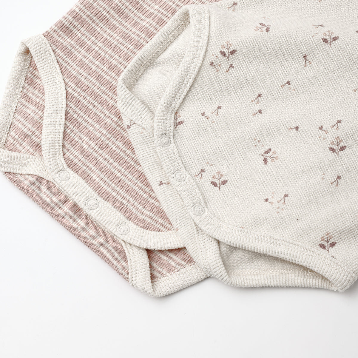 Two soft baby bodysuit fabrics laid flat on a white background: one with thin dusty pink and cream stripes, and one in warm off-white with small scattered floral motifs.