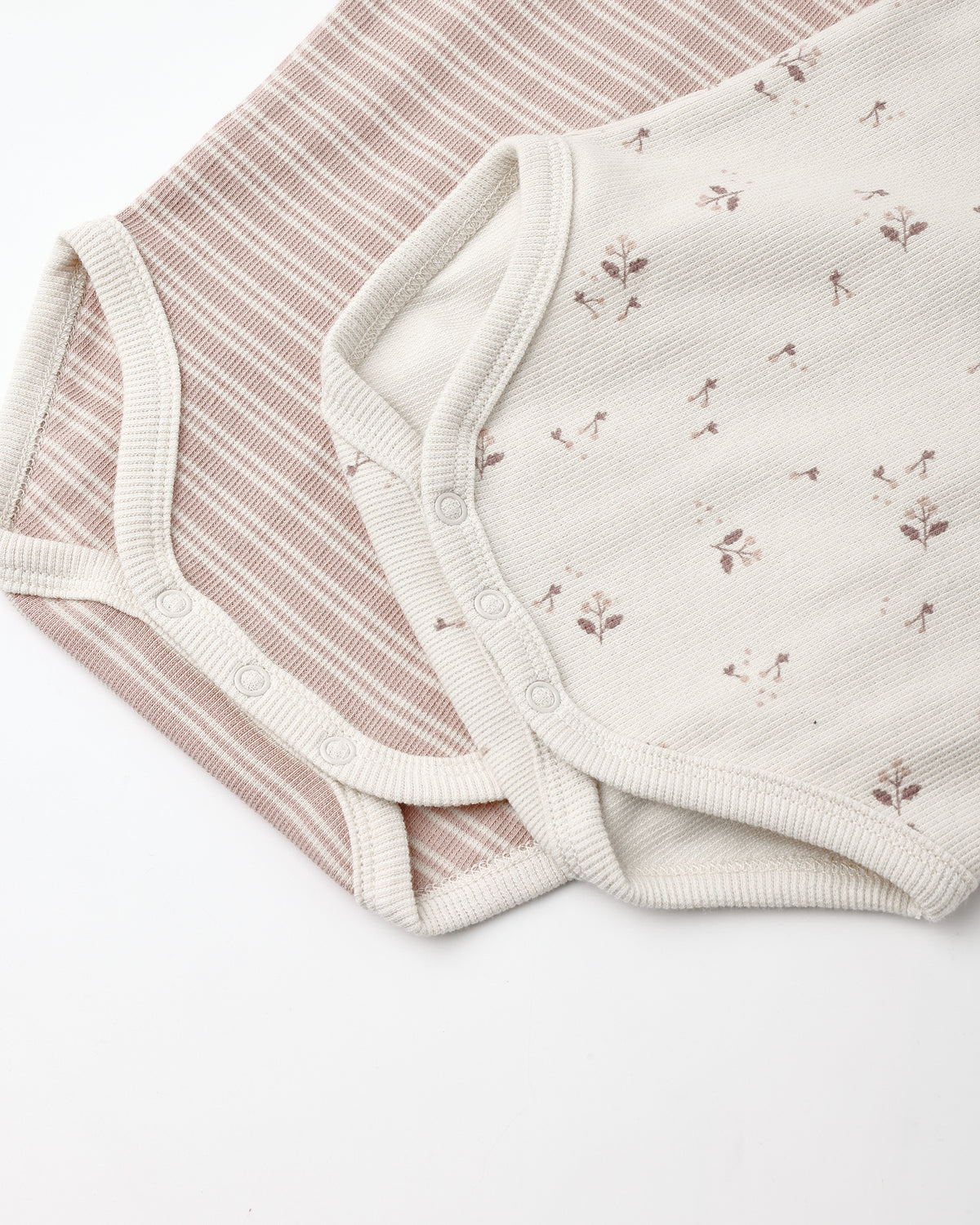 Two soft baby bodysuit fabrics laid flat on a white background: one with thin dusty pink and cream stripes, and one in warm off-white with small scattered floral motifs.