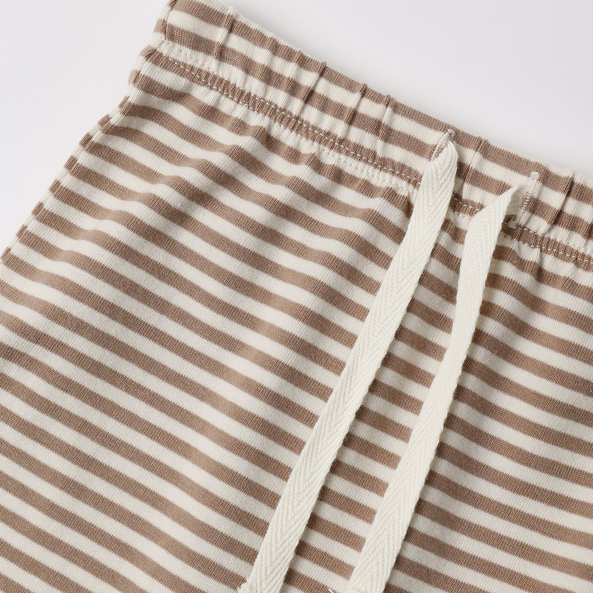 Close-up of striped drawstring shorts with a beige and off-white horizontal stripe pattern and matching off-white drawstrings, shown on a light gray background.