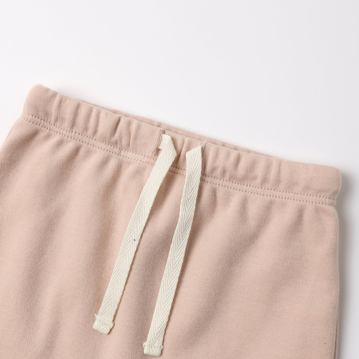 Close-up of a pair of soft blush pink sweatpants with an off-white drawstring, shown against a plain light background.