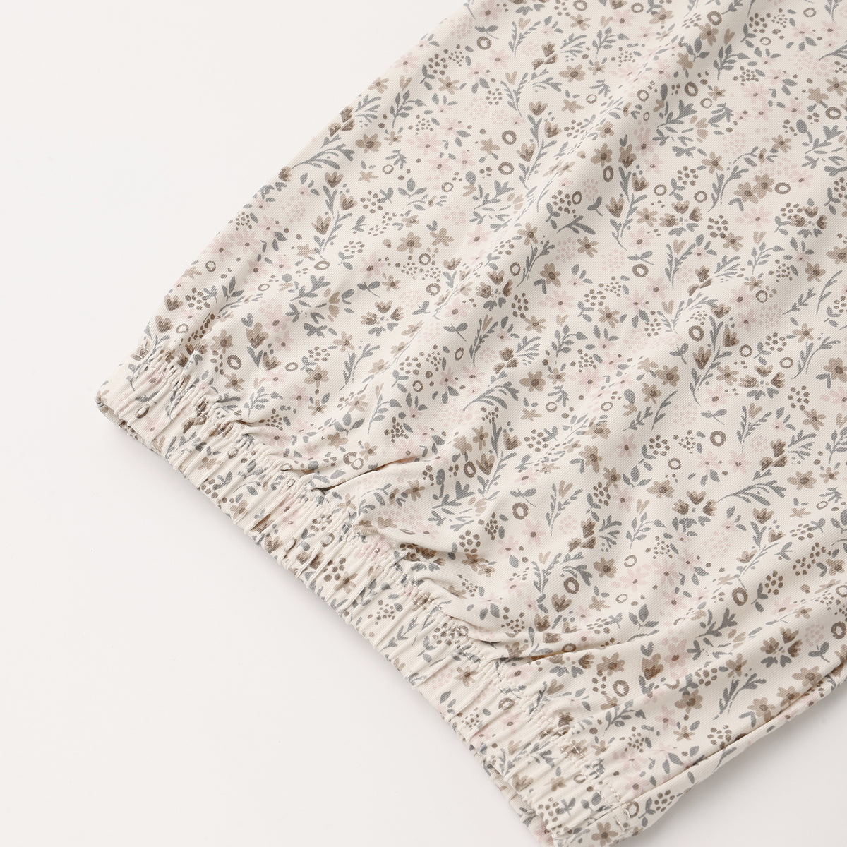 A close-up of a lightweight fabric with a delicate, small-scale floral and botanical print in muted gray and brown tones on a warm cream background, finished with an elasticized ruffled hem.