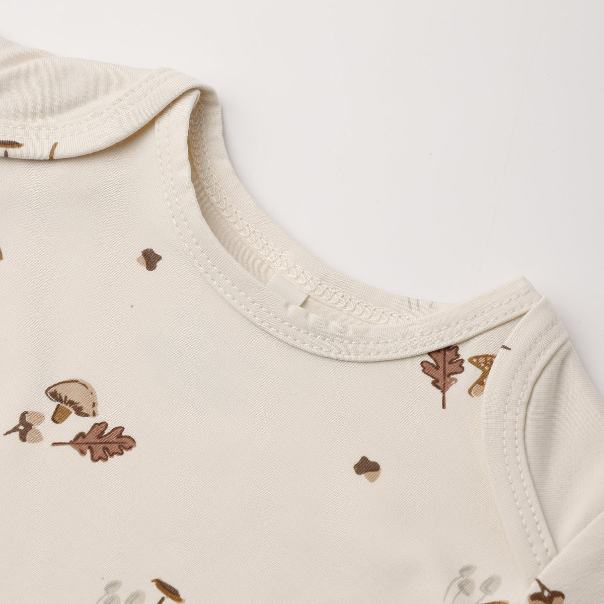 Close-up of a cream-colored baby top with a delicate brown and beige autumn leaf and mushroom print, showing the ribbed collar and shoulder seams.
