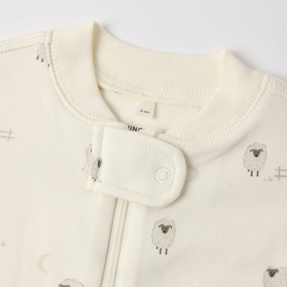 Close-up of a baby garment in warm off-white/cream with a small sheep print and a snap-button closure at the neckline.