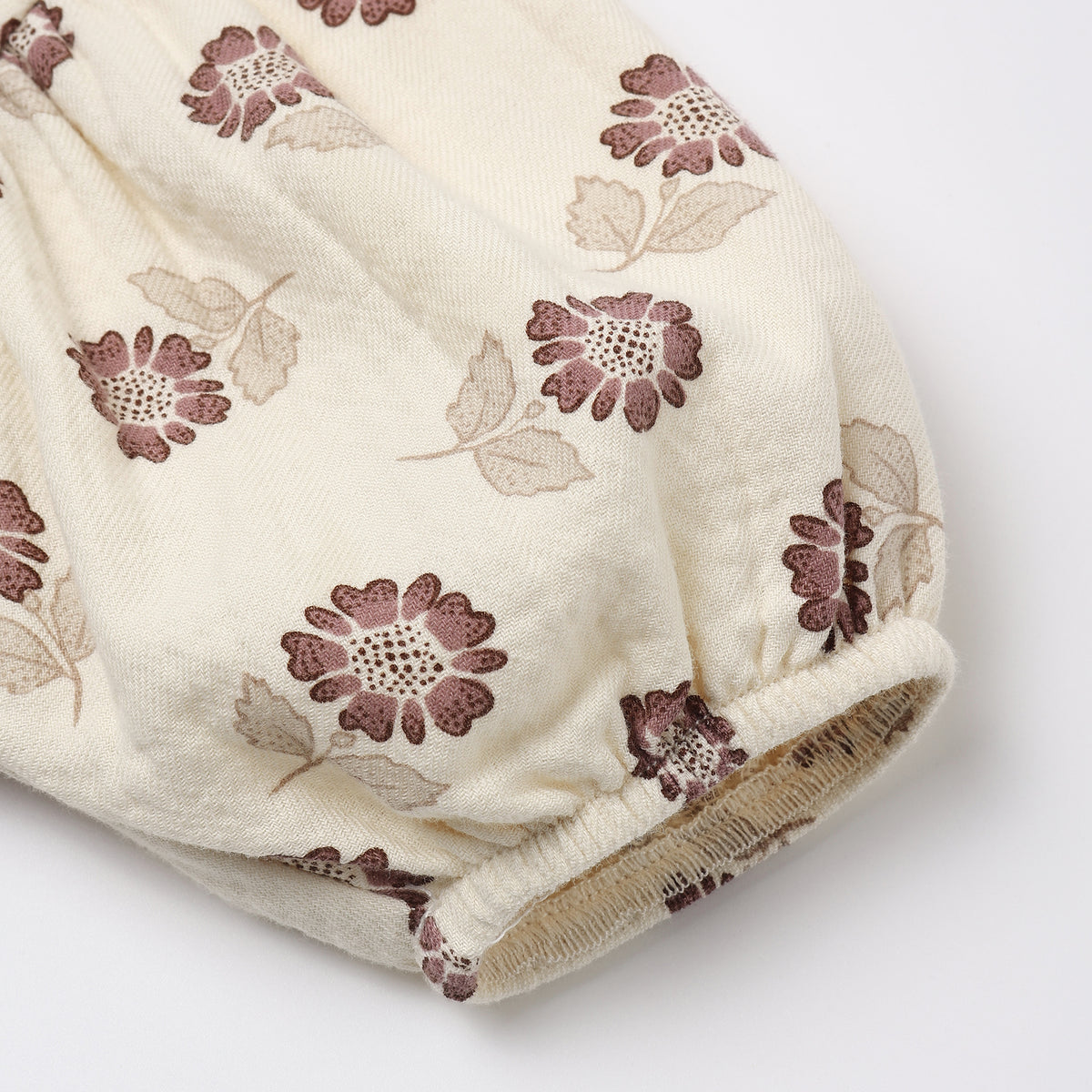 A close-up of a cream-colored fabric garment, likely a baby romper or skirt, featuring an all-over floral print in dusty mauve and beige, finished with a gathered elastic cuff.