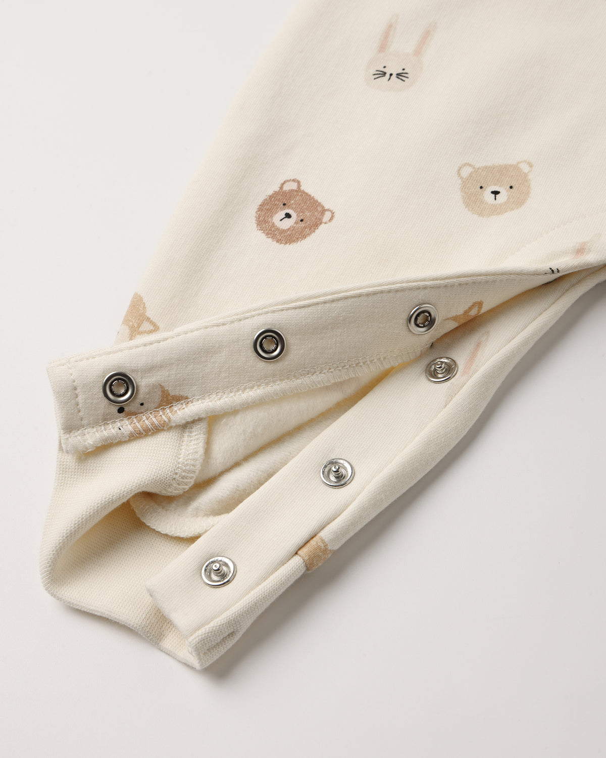 Close-up of a cream-colored baby garment with embroidered teddy bear and bunny faces, featuring silver metal snap buttons along the folded cuff.