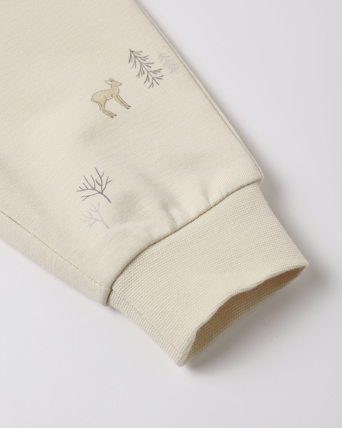 Close-up of a cream-colored sweatshirt sleeve featuring a small embroidered deer and delicate tree motifs near the ribbed cuff.