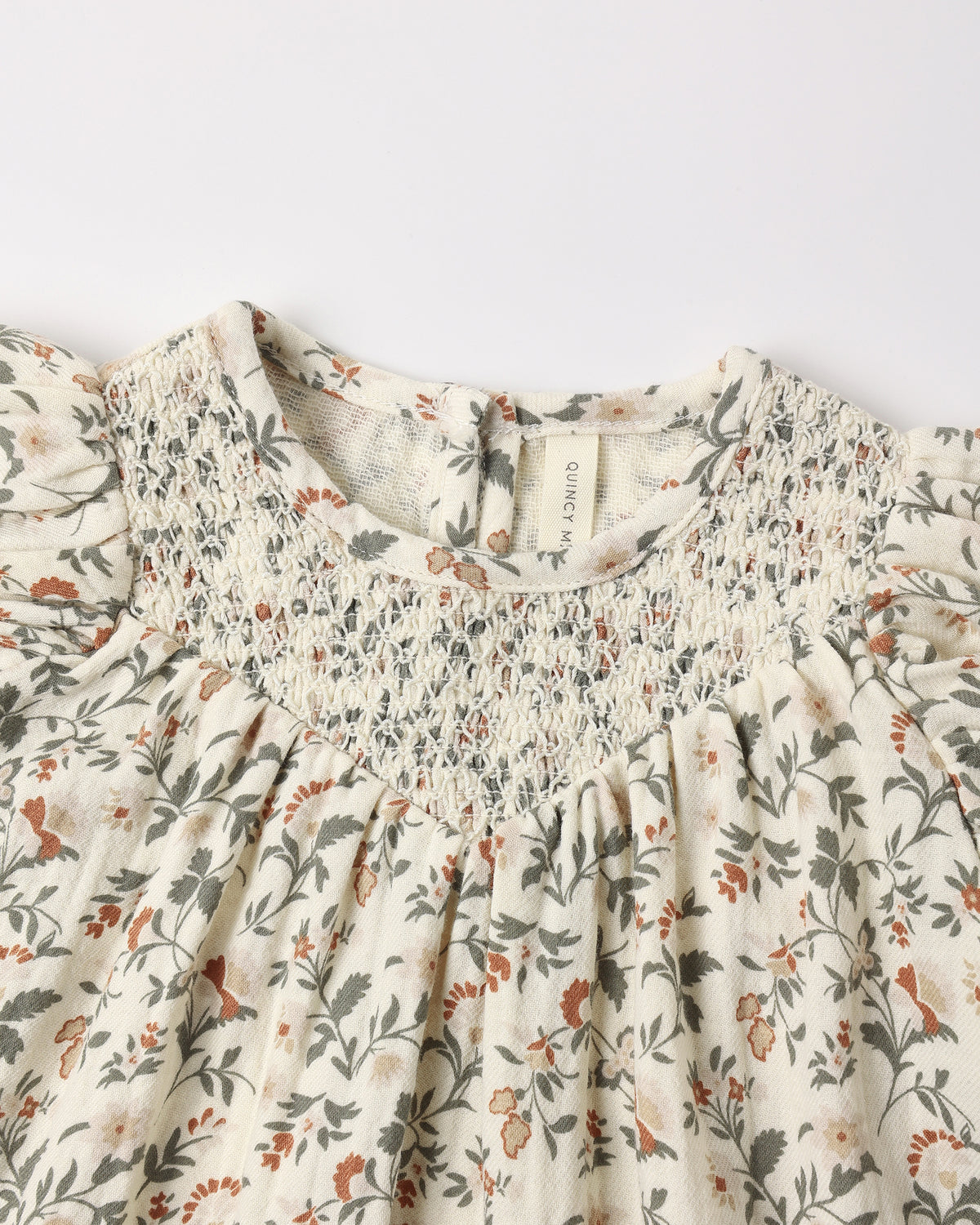 Carina Dress - Holiday Floral by Quincy Mae shown in a close-up highlighting the natural fabric, festive floral print, short bubble sleeves, and diamond smocking around the neckline.