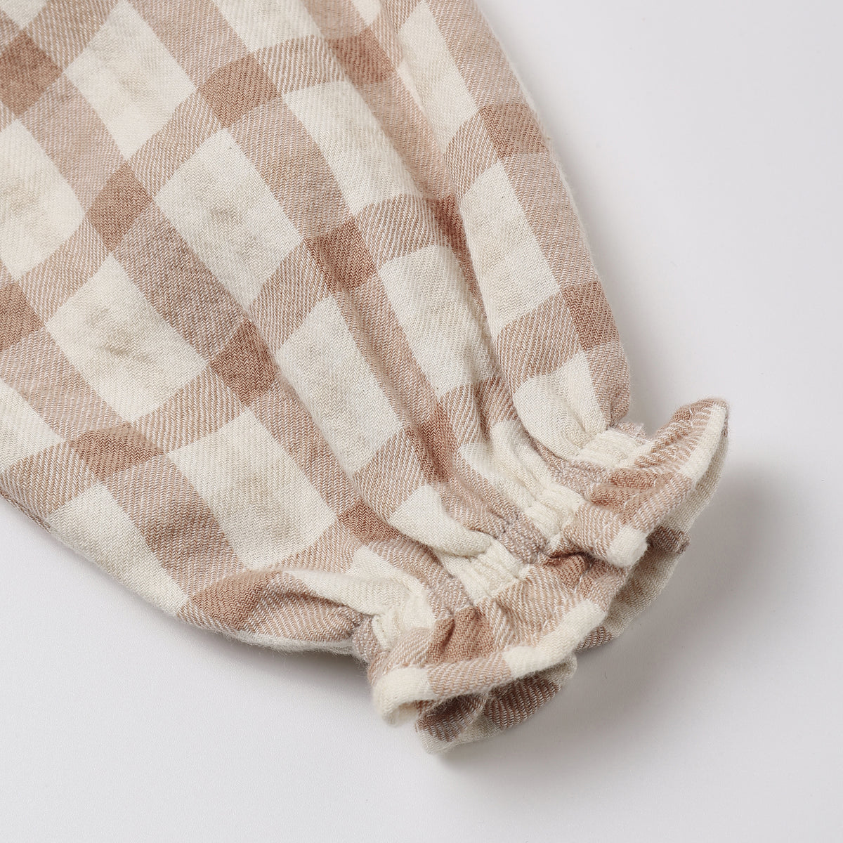 A close-up of a soft cotton fabric sleeve or cuff with a beige and off-white gingham check pattern and a gathered elastic ruffle at the end, shown on a light gray background.