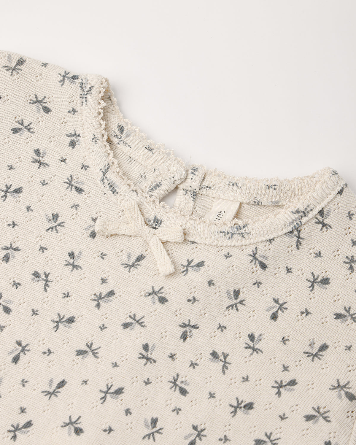 A cream-colored children's pointelle top featuring an all-over small floral print and a delicate bow detail at the neckline