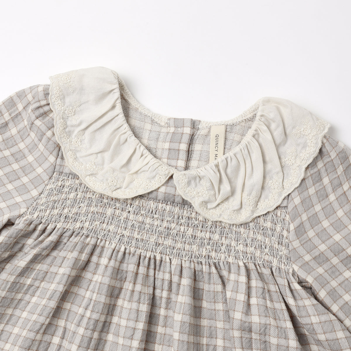 Close-up of a children's dress with a gray and white gingham pattern, featuring a smocked bodice and a large cream-colored ruffled lace Peter Pan collar.