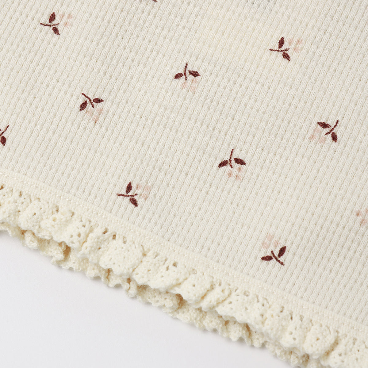 Close-up of a cream-colored ribbed knit fabric with a delicate ruffled lace trim and small embroidered floral motifs in brown and soft pink.