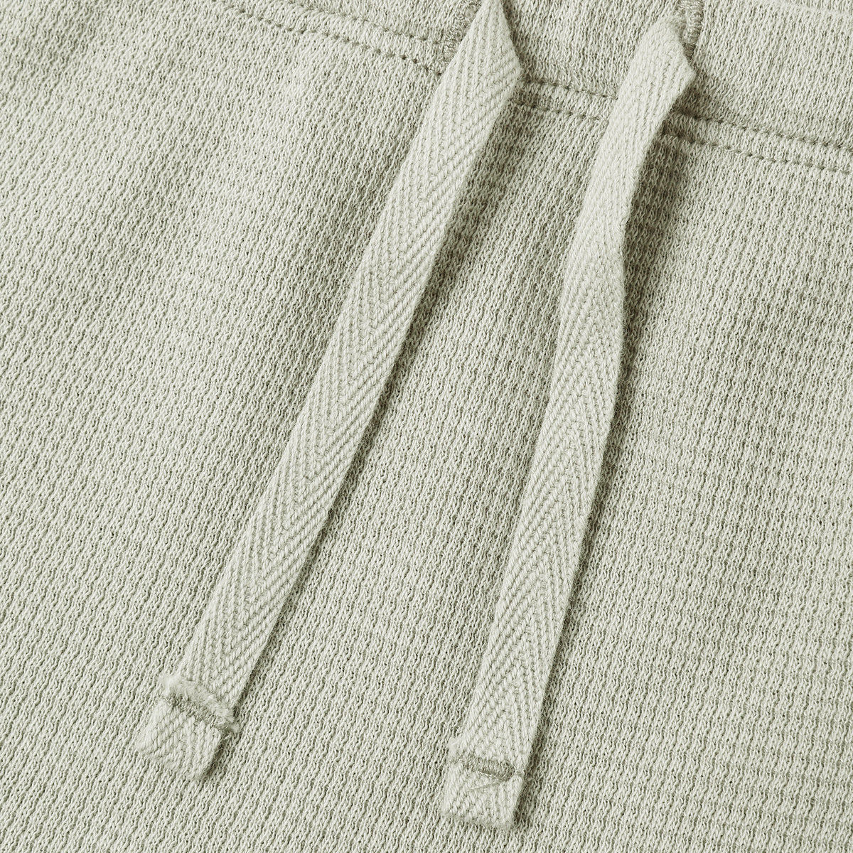 Close-up of a light sage green textured knit garment, showing two matching drawstrings against a white background.