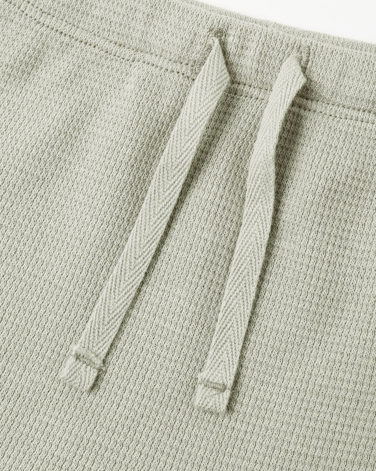 Close-up of a light sage green textured knit garment, showing two matching drawstrings against a white background.