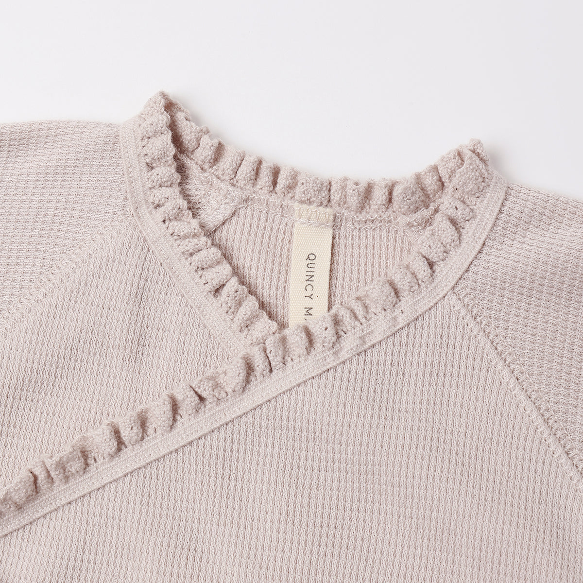 Close-up of a knit sweater in a light warm beige or taupe color, showing a waffle-knit texture, a ruffled collar, and a cream-colored interior neck label that reads 'QUINCY ART'.