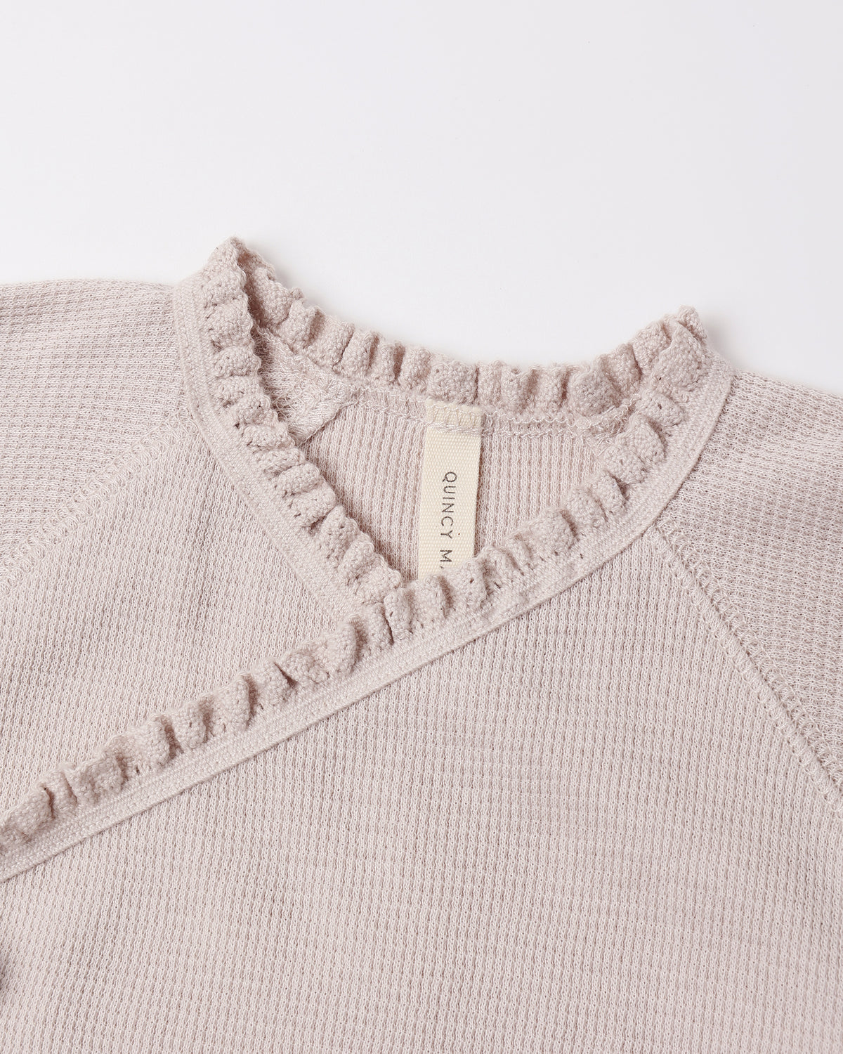 Close-up of a knit sweater in a light warm beige or taupe color, showing a waffle-knit texture, a ruffled collar, and a cream-colored interior neck label that reads 'QUINCY ART'.