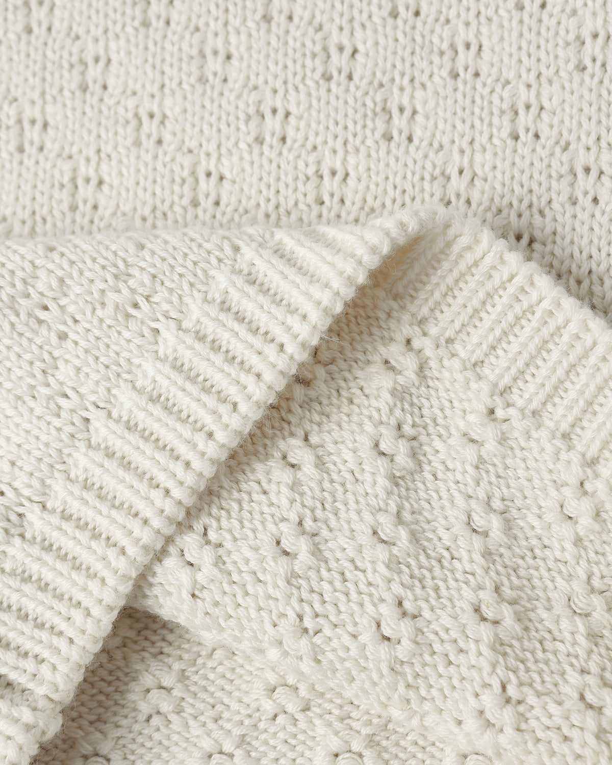 Close-up of a cream-colored knit fabric with a textured bobble pattern and ribbed trim, folded over to show the material detail.