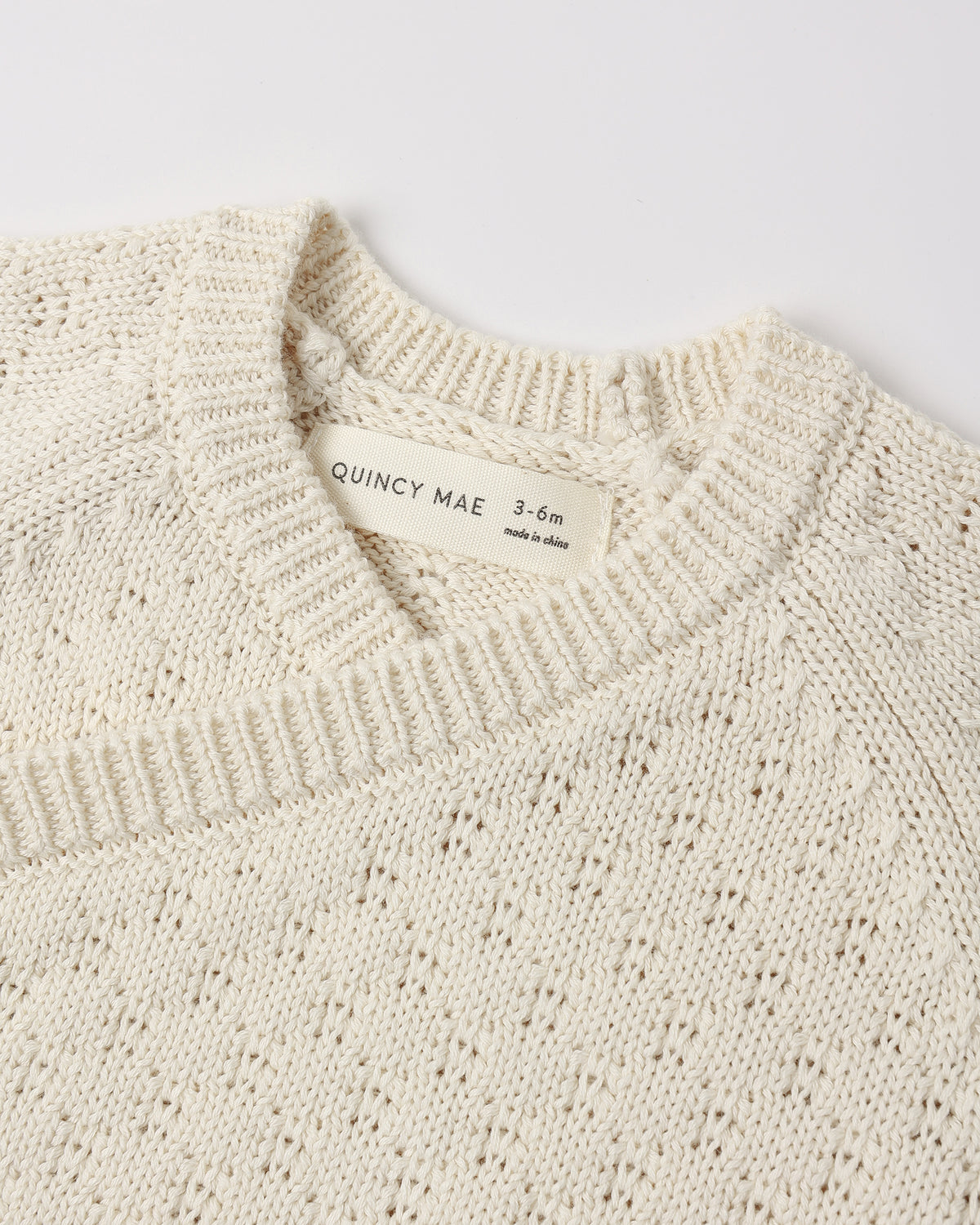 Close-up of a cream-colored knit sweater with a visible neck label reading 'QUINCY MAE 3-6m made in Spain'.