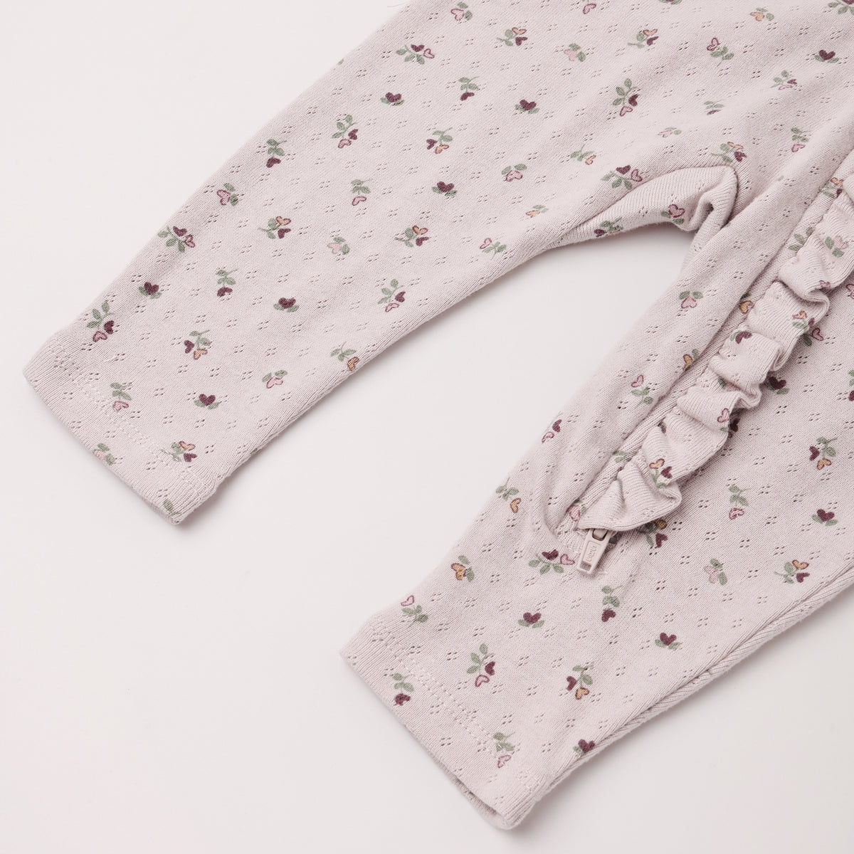 Close-up of a baby or toddler long-sleeve romper or pajama top in a pale blush pink eyelet fabric with a small scattered floral print, featuring a ruffled trim detail on the side.