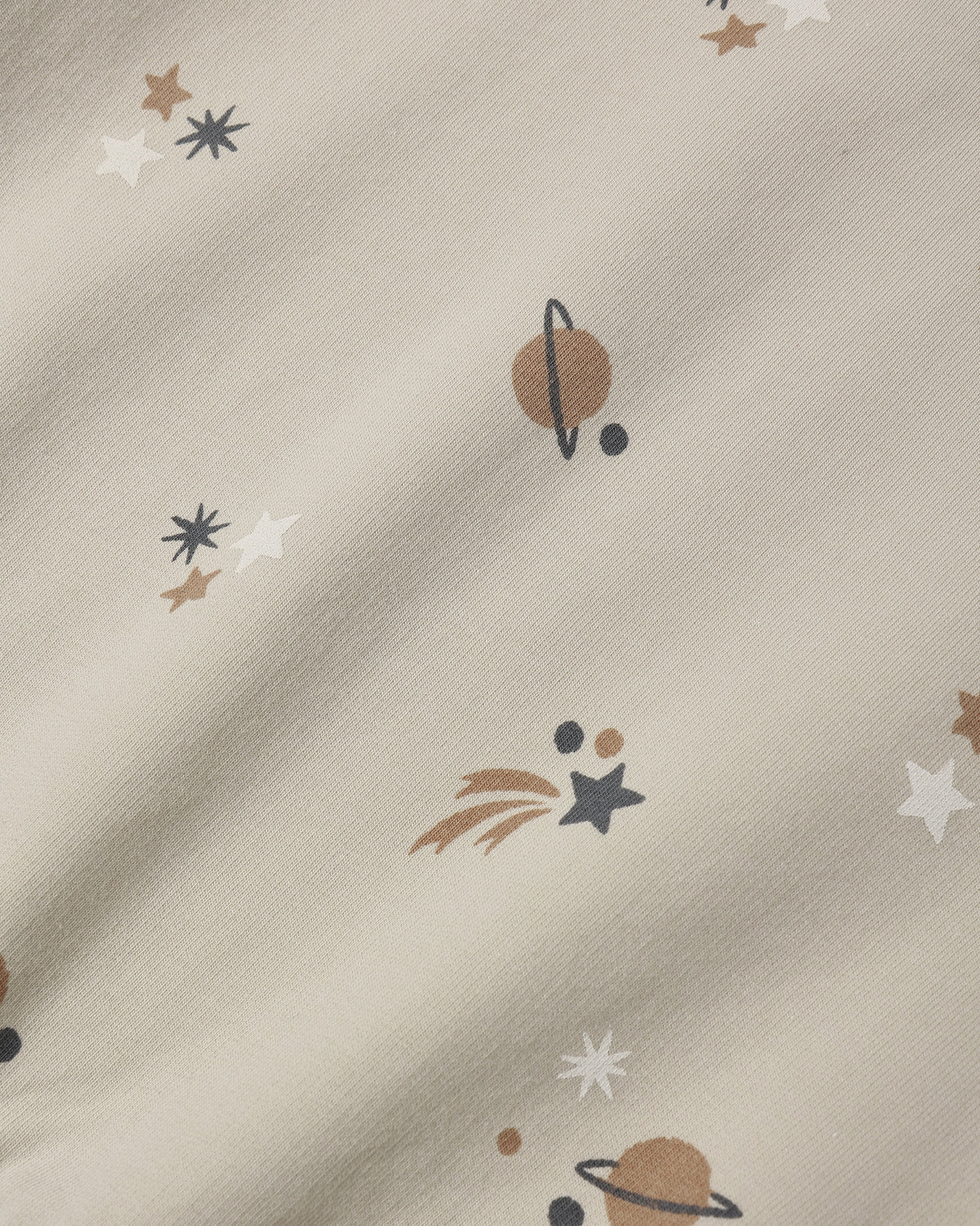 A close-up of a cream-colored fabric with a printed space theme, featuring planets, stars, and shooting stars in brown, white, and dark gray.