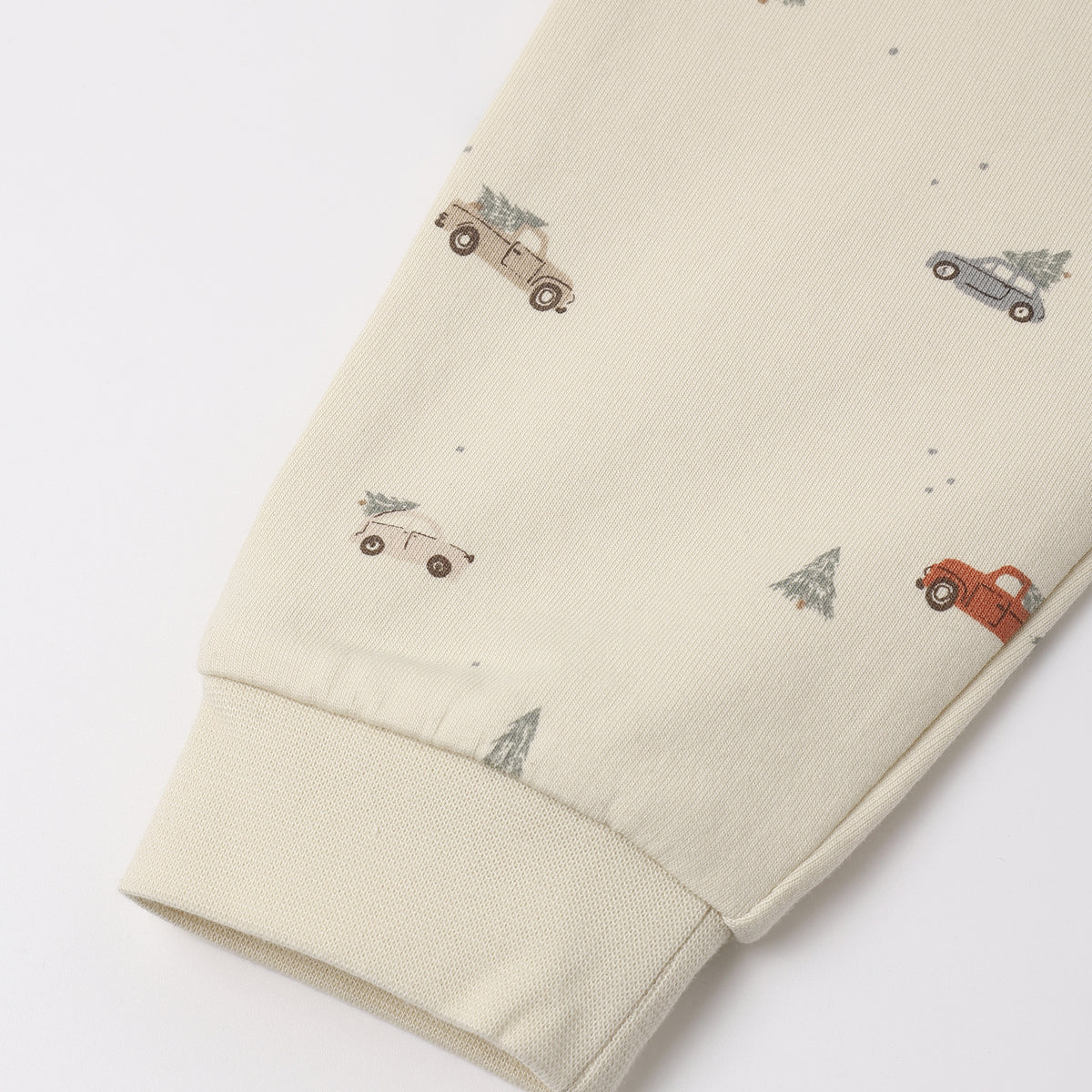 Close-up of cream-colored organic cotton fabric featuring a "Holiday Drive" print with tiny vintage cars and pine trees, showing a ribbed sleeve cuff.