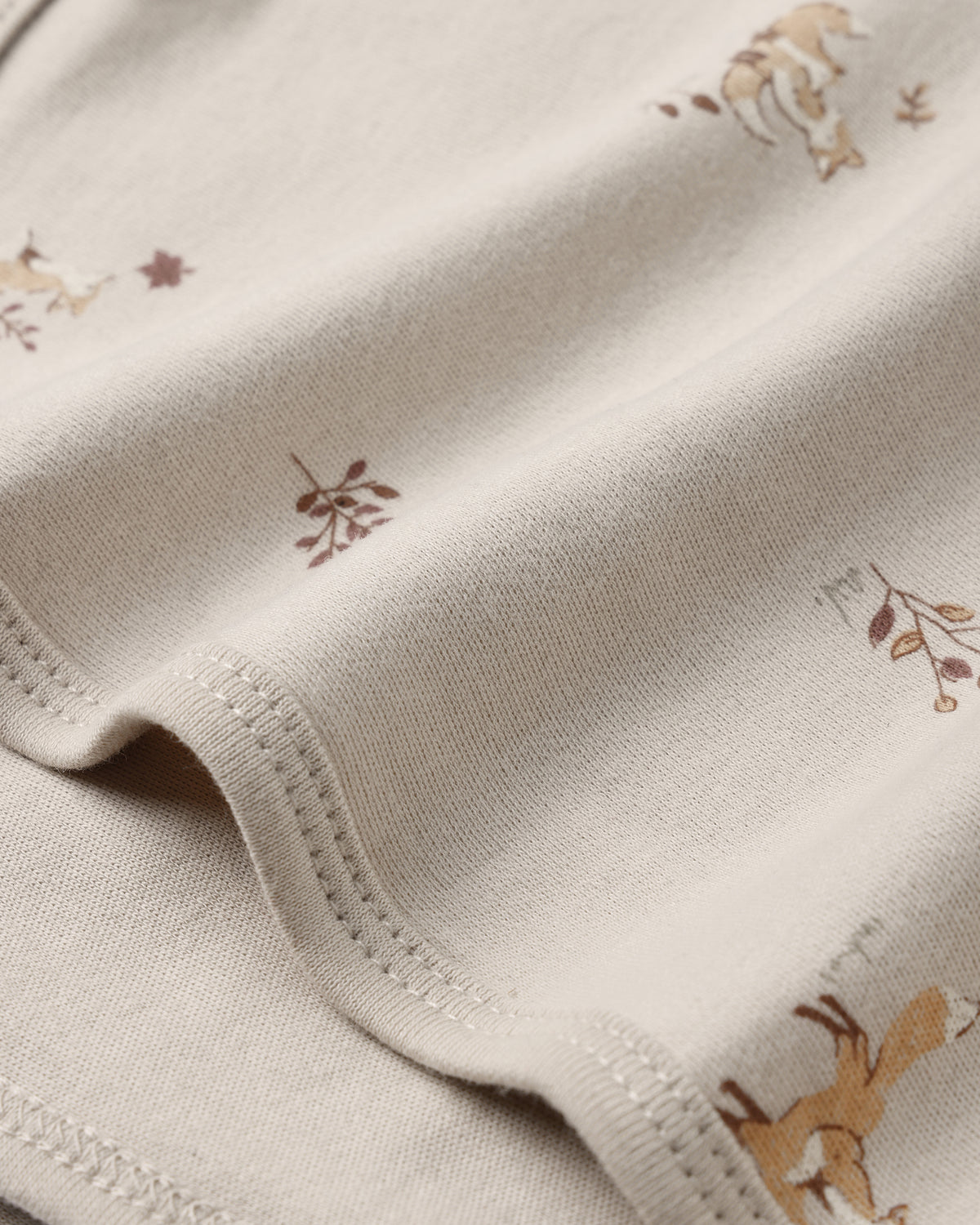 Close-up of a cream-colored knit fabric with a delicate print of small brown and tan woodland animals and leaves, showing the fine knit texture and neat stitching.