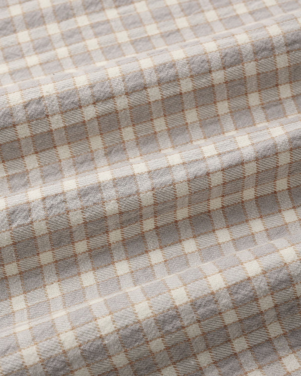A close-up of a woven fabric with a soft plaid pattern in light gray, cream, and thin tan lines, showing the texture and folds of the material.