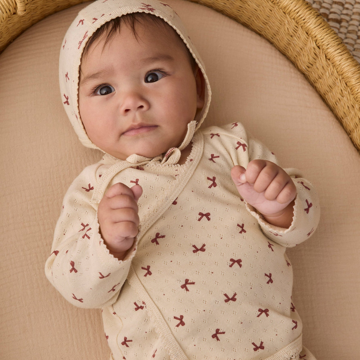 An infant wearing the Quincy Mae red bows print long-sleeve top and matching bonnet, lying comfortably in a woven basket.