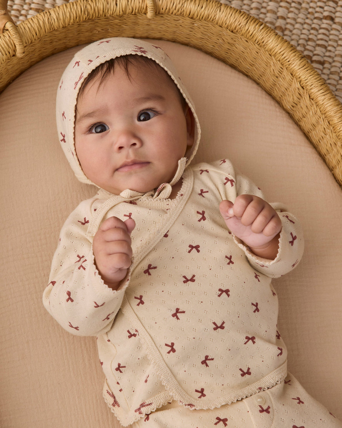 An infant wearing the Quincy Mae red bows print long-sleeve top and matching bonnet, lying comfortably in a woven basket.