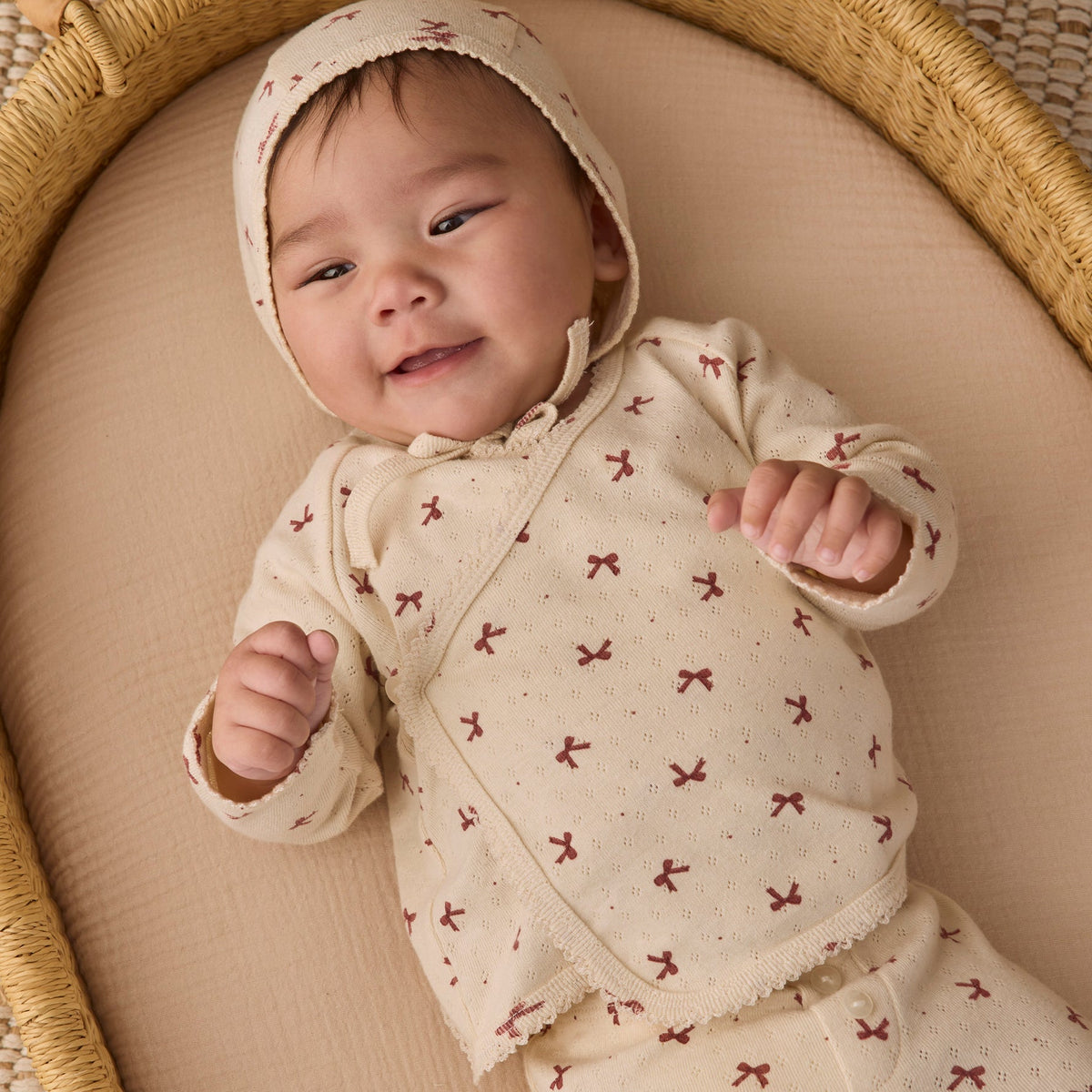 An infant wearing the Quincy Mae red bows print take-home set, featuring a matching bonnet and long-sleeve top, lying comfortably in a woven basket.