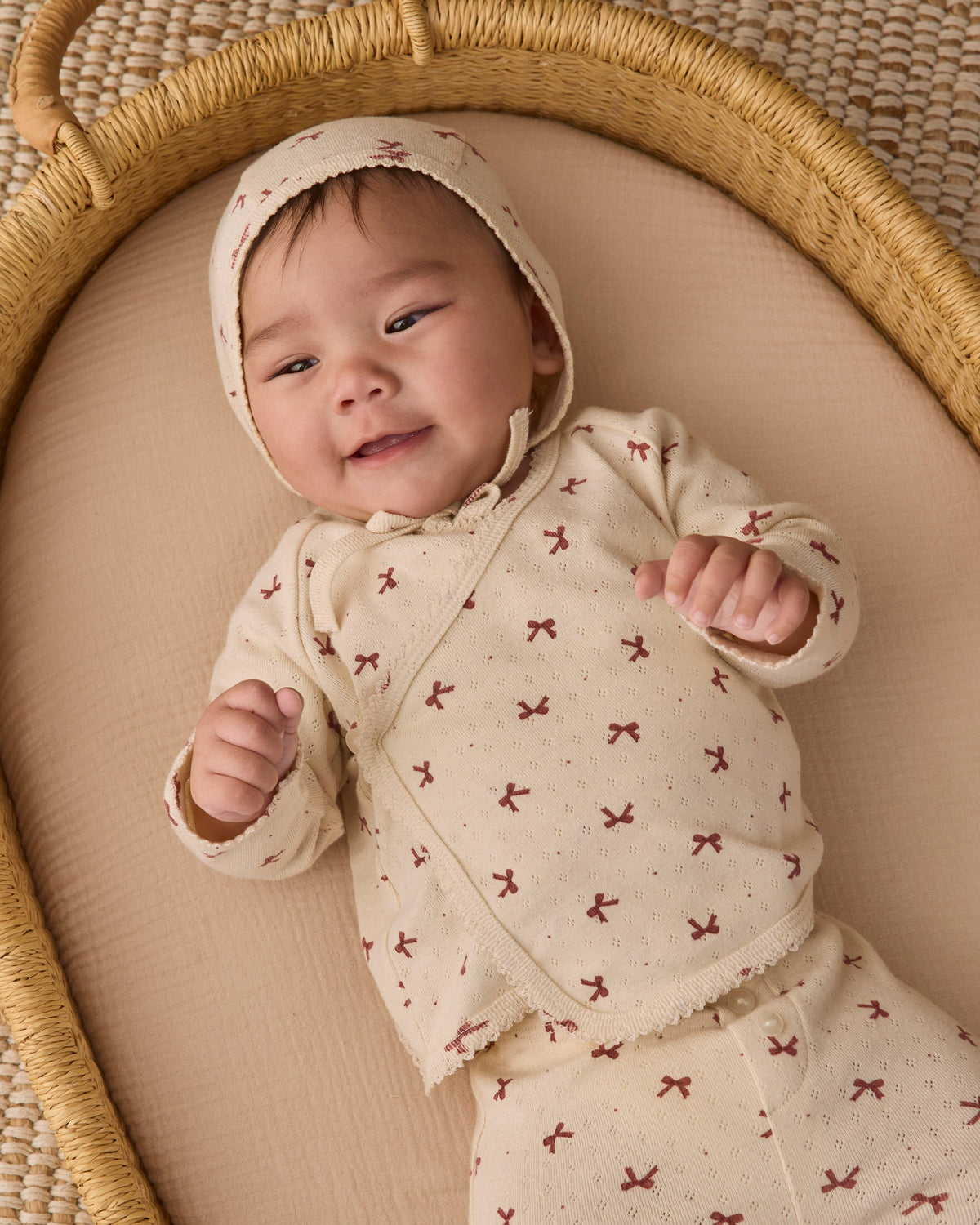 An infant wearing the Quincy Mae red bows print take-home set, featuring a matching bonnet and long-sleeve top, lying comfortably in a woven basket.