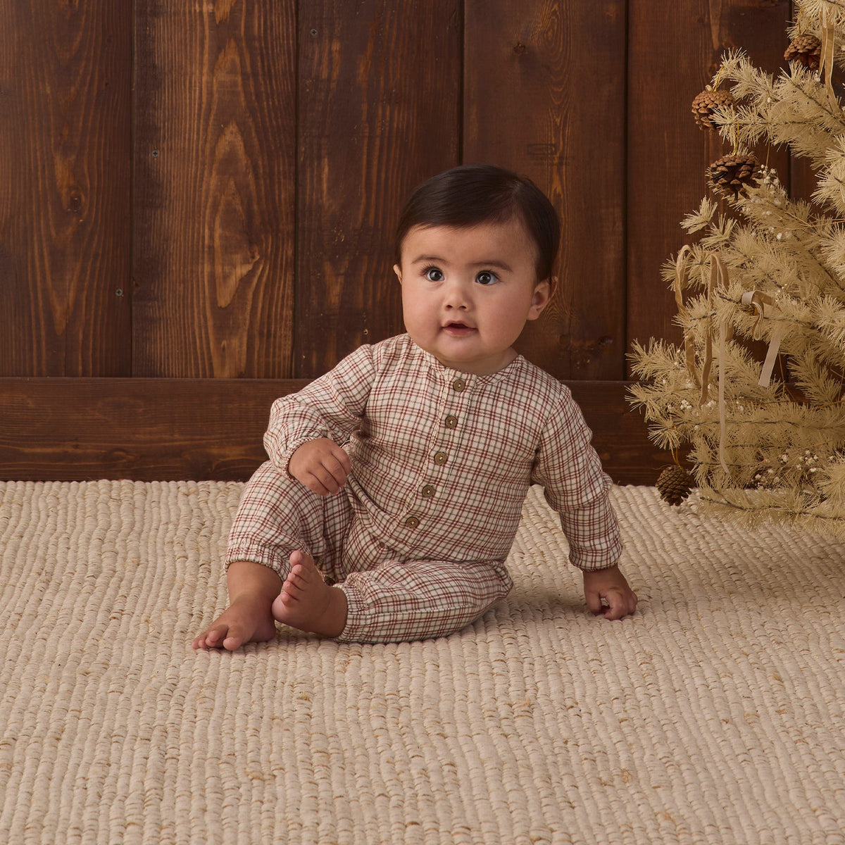 A baby crawling on a textured cream rug in front of a wood-paneled wall, wearing a long-sleeve Christmas plaid jumpsuit.