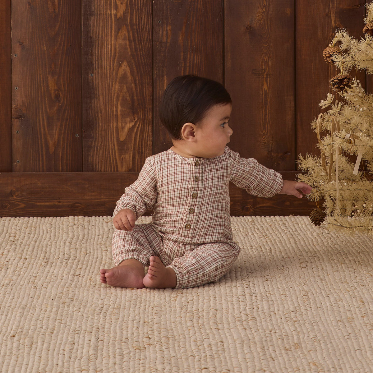 A baby sitting on a textured cream rug in front of a wood-paneled wall, wearing a long-sleeve Christmas plaid jumpsuit with a button placket on the chest.
